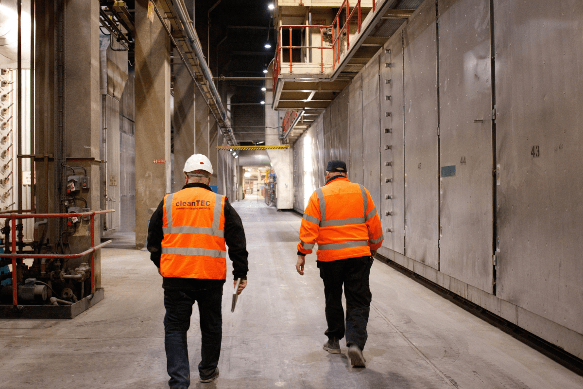 Two workers in high-visibility orange jackets, one with a hard hat and a CleanTEC logo on the back, walking through an industrial facility with tall concrete walls and overhead structures.